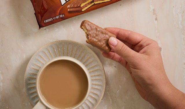 A cup of tea on a table and a hand holding a Tim Tam biscuit